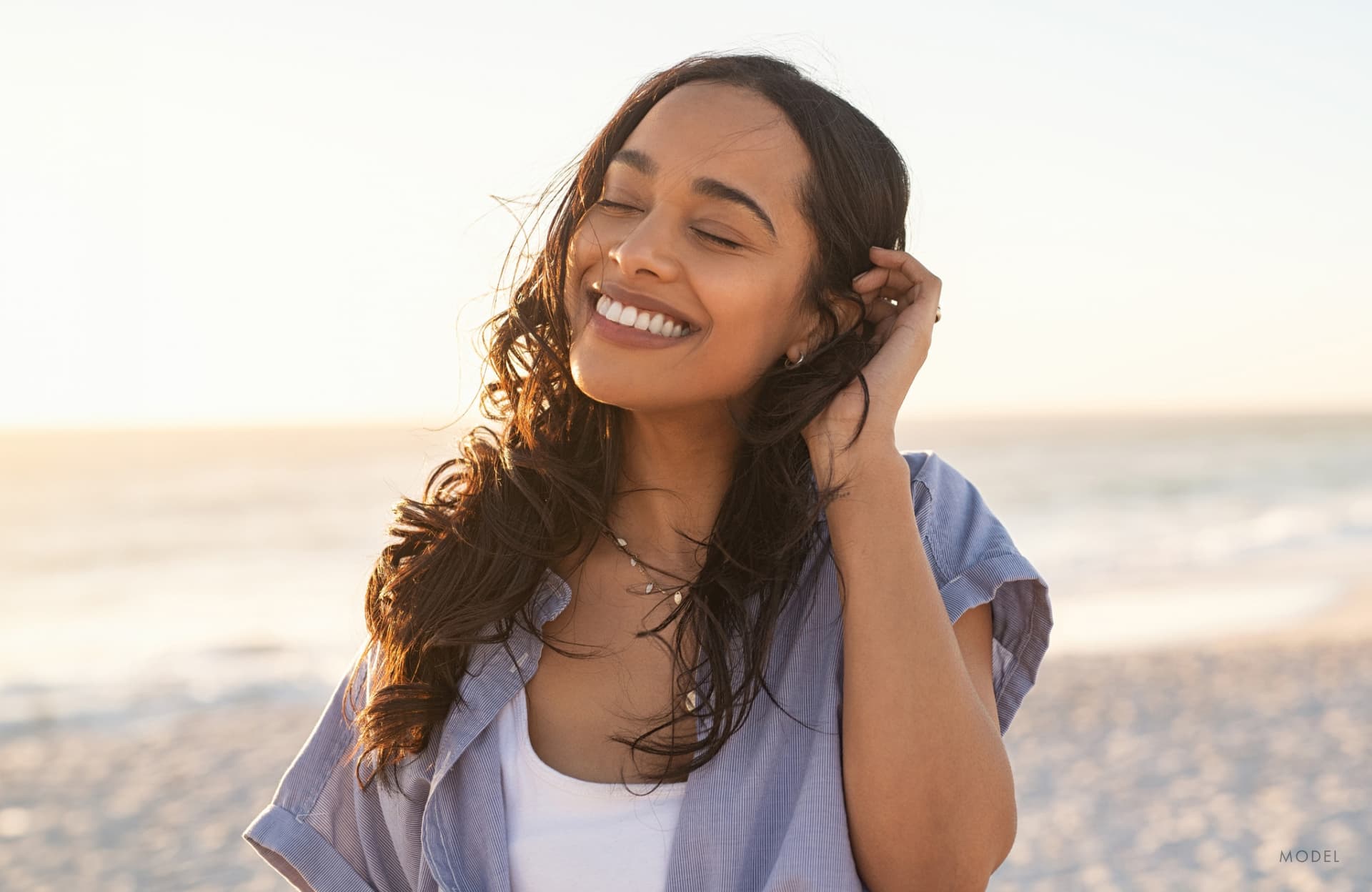 A model image of a woman smiling with her eyes closed while standing on the beach.